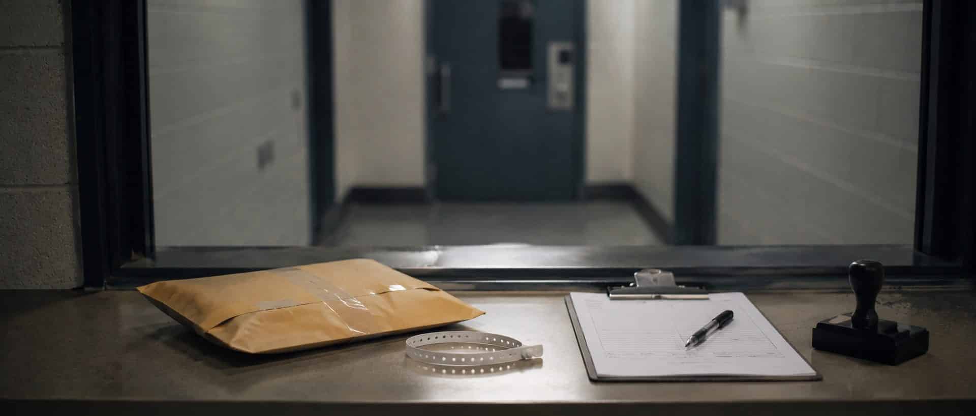A county jail property counter with a sealed property envelope, wristband and release paperwork, and a closed steel door beyond.